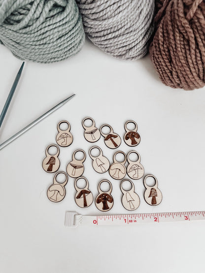 A total of 14 wood stitch markers with a variety of hand-drawn mushroom designs are displayed next to a measuring tape that shows they&