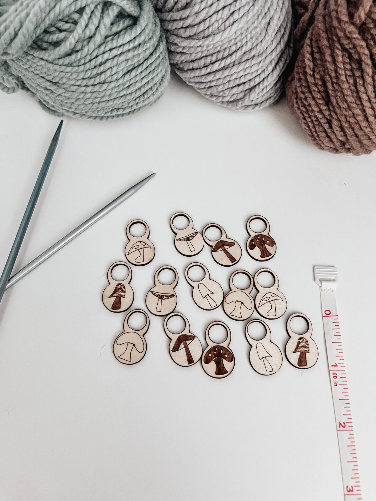 A total of 14 wood stitch markers with a variety of hand-drawn mushroom designs are displayed next to a measuring tape that shows they&