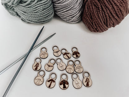 A total of 14 wood stitch markers with a variety of hand-drawn mushroom designs are displayed with a pair of metal knitting needles and three skeins of merino yarn are in the background.
