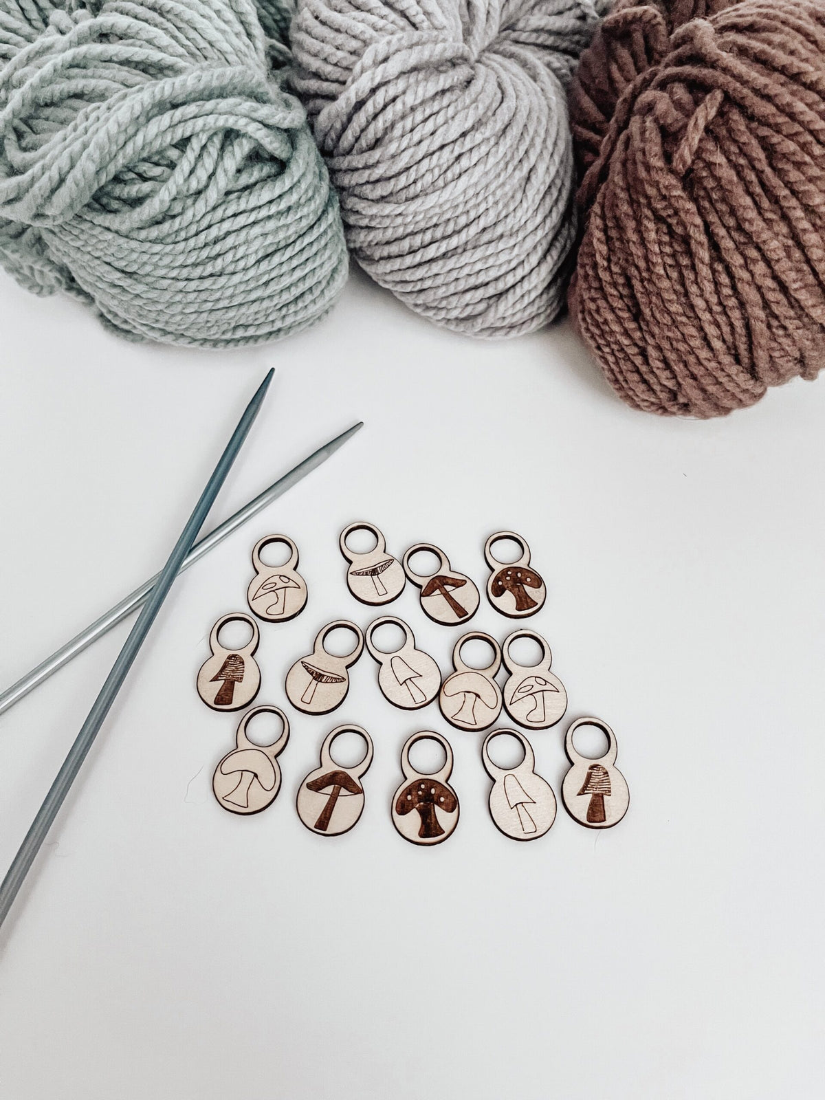A total of 14 wood stitch markers with a variety of hand-drawn mushroom designs are displayed with a pair of metal knitting needles and three skeins of merino yarn are in the background.