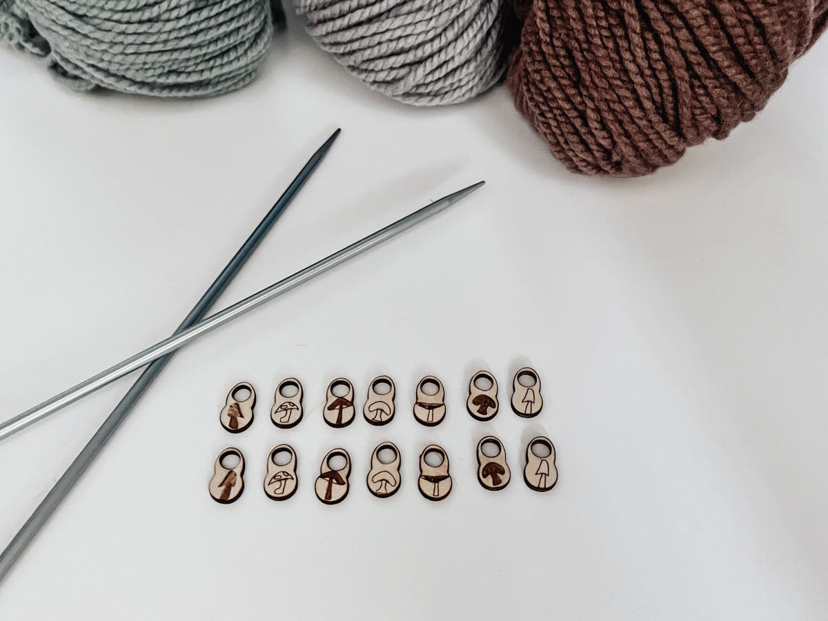 A total of 14 wood stitch markers with a variety of hand-drawn mushroom designs are displayed with a pair of metal knitting needles and three skeins of merino yarn are in the background.