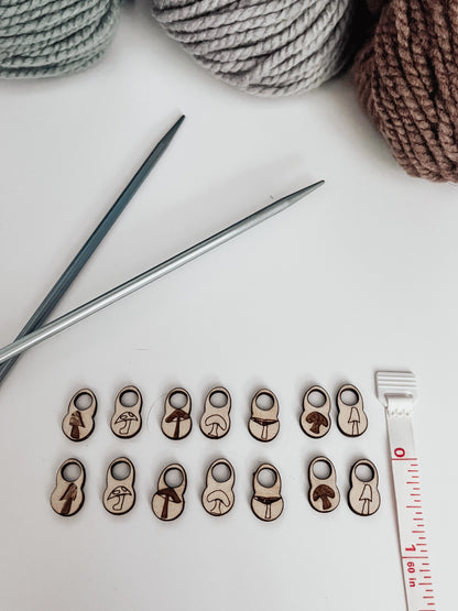 A total of 14 wood stitch markers with a variety of hand-drawn mushroom designs are displayed next to a measuring tape that shows they&