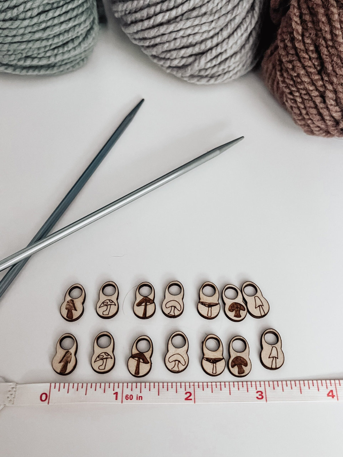 A total of 14 wood stitch markers with a variety of hand-drawn mushroom designs are displayed next to a measuring tape that shows they&