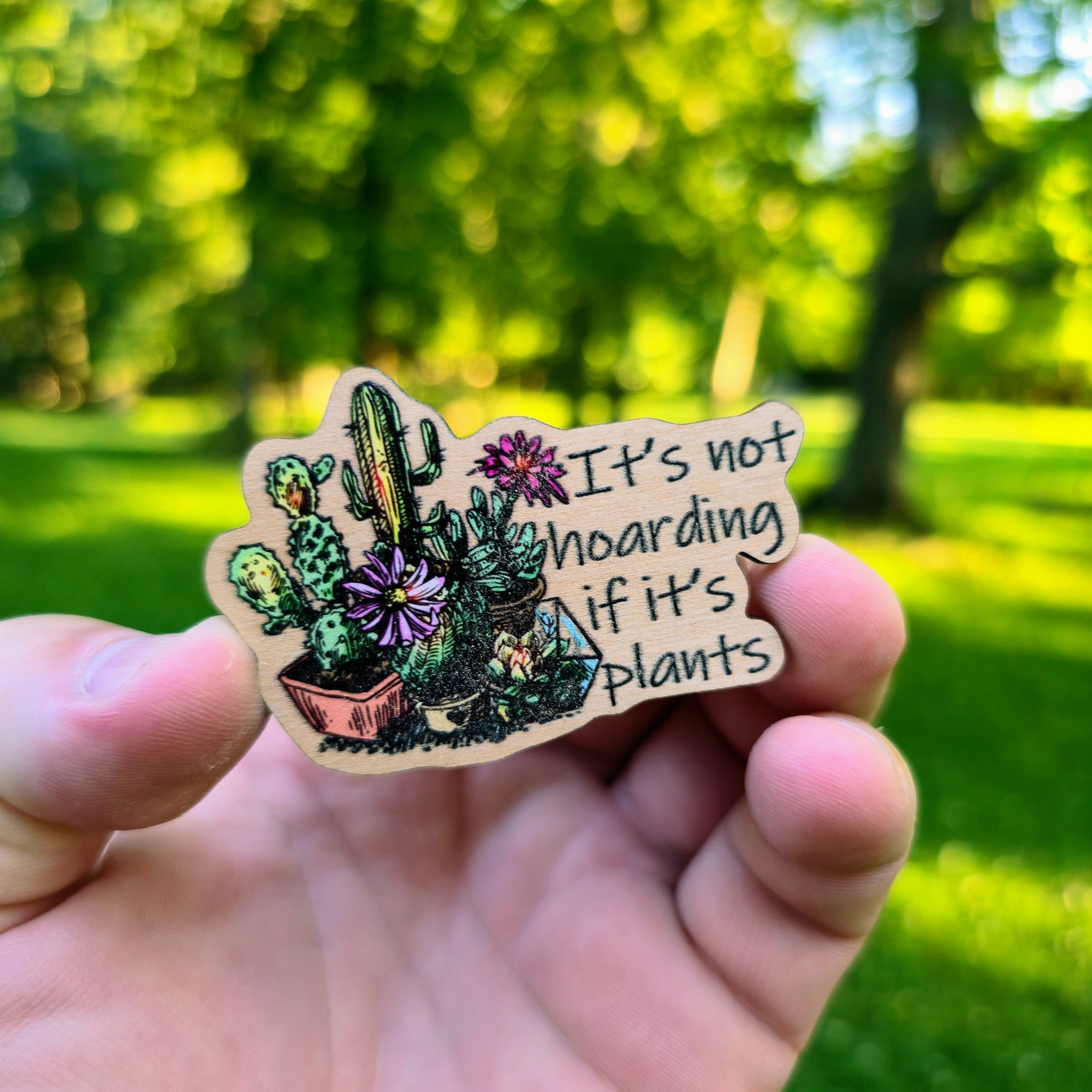 Hand holding a wooden magnet with plant illustration and text against a blurred green outdoor background