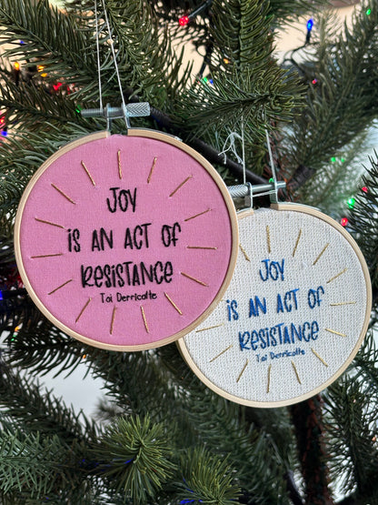 Two hand embroidery hoops that read, "Joy is an act of resistance" - Toi Derricotte. Hoops are hanging on a Christmas tree as Christmas ornaments. Text is a handwriting font. One ornament is hand embroidered in black embroidery thread. Words are centered in a burst of metallic gold rays. Fabric is light pink cotton. Framed in light wood embroidery hoop. In a second hoop with the same message, the words are sewn in blue thread with gold metallic rays on a sparking cream linen fabric.
