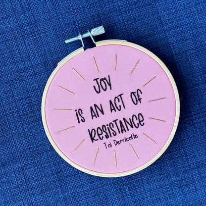 "Joy is an act of resistance" - Toi Derricotte. Text is a handwriting font and hand embroidered in black embroidery thread. Words are centered in a burst of golden, metallic rays. Fabric is light pink cotton. Framed as wall art or christmas ornament in light wood embroidery hoop.