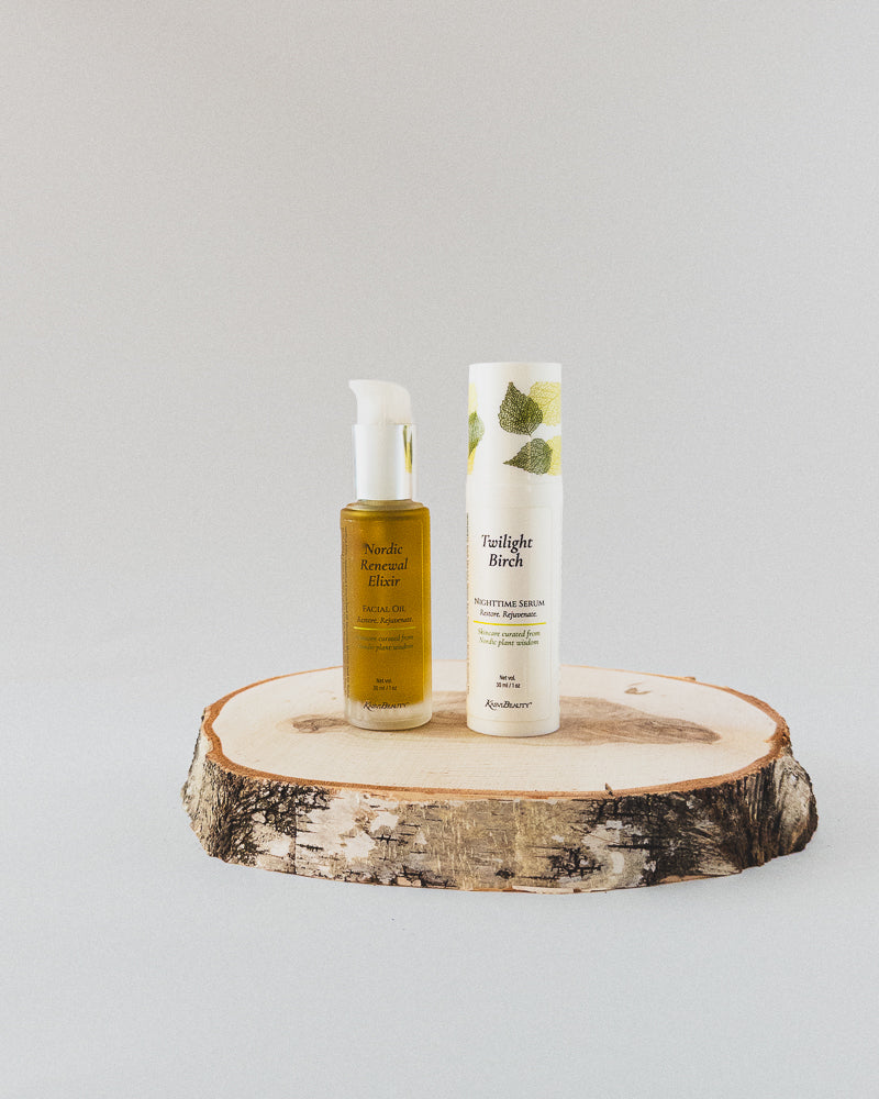 Two skincare products from the brand Nordic Renewal Elixir Facial Oil with a white pump, and a white airless pump of Twilight Birch Nighttime Renewal Serum with green leaf graphics, displayed on a round birch wood slice against a light gray background.