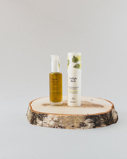 Two skincare products from the brand Nordic Renewal Elixir Facial Oil with a white pump, and a white airless pump of Twilight Birch Nighttime Renewal Serum with green leaf graphics, displayed on a round birch wood slice against a light gray background.
