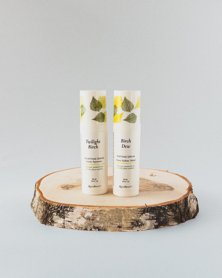 Two white airless bottles standing upright on a round natural wood slice coaster against a light gray studio background. The tubes have green leaf illustrations near the top and readable text including &