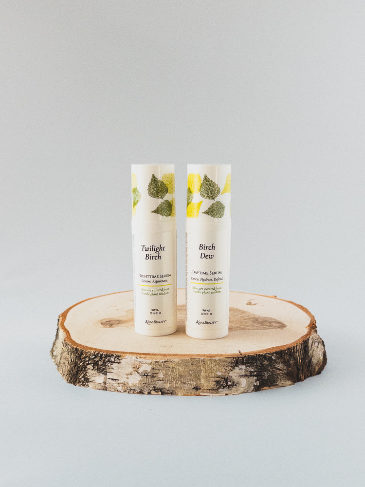Two white airless bottles standing upright on a round natural wood slice coaster against a light gray studio background. The tubes have green leaf illustrations near the top and readable text including &