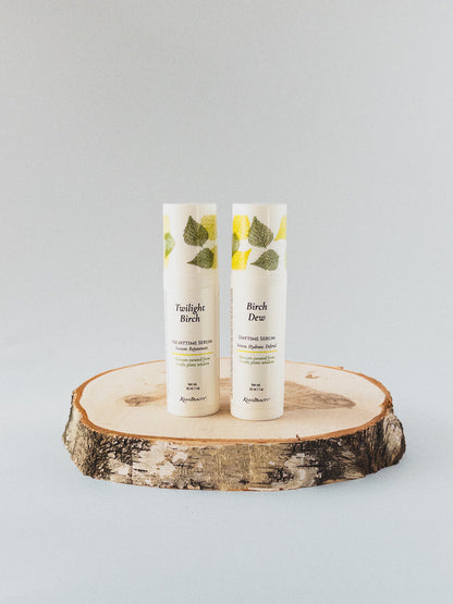 Two white airless bottles standing upright on a round natural wood slice coaster against a light gray studio background. The tubes have green leaf illustrations near the top and readable text including &
