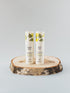 Two white airless bottles standing upright on a round natural wood slice coaster against a light gray studio background. The tubes have green leaf illustrations near the top and readable text including &