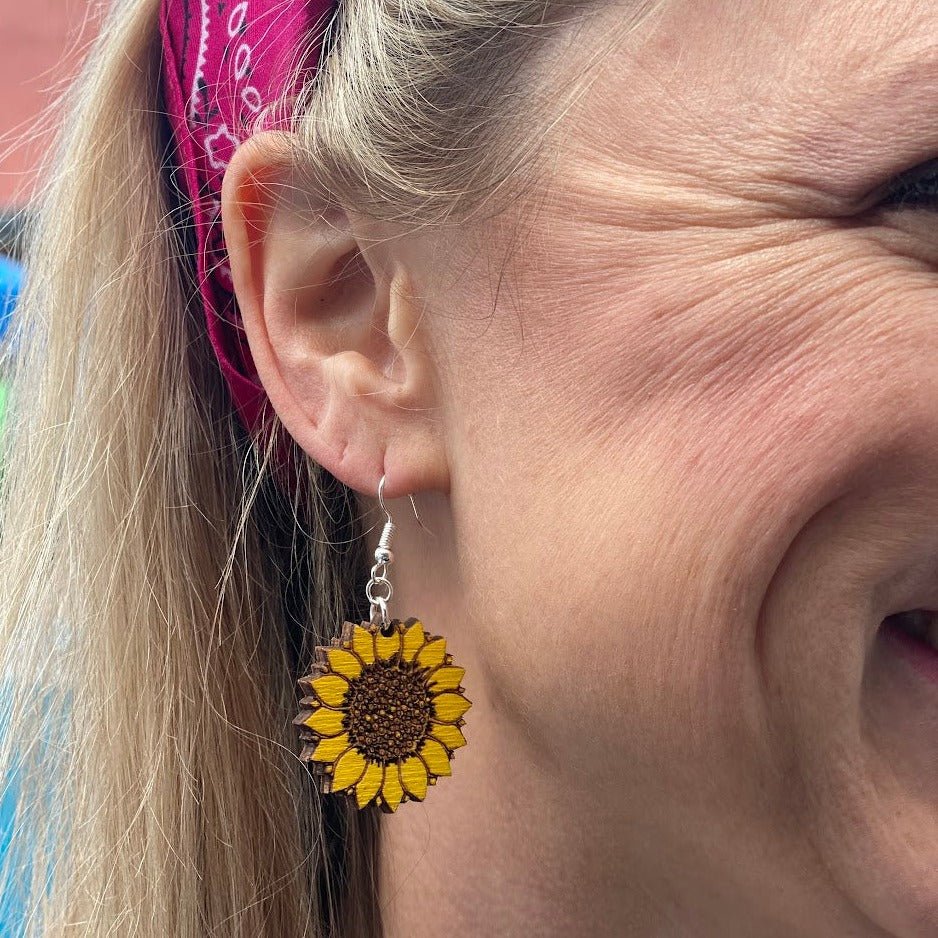 Close-up of a hand-painted wooden sunflower dangle earring worn on the ear, featuring vibrant yellow petals and a detailed brown sunflower center. This detailed image highlights the handcrafted artwork and lightweight design with a hypoallergenic .925 sterling silver hook, making these earrings perfect for gardeners, flower lovers, farmhouse style, and everyday wear.