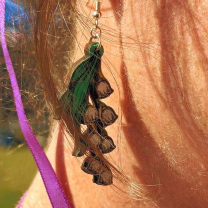 Close-up of hand-painted wooden lily of the valley dangle earrings with green leaves and white bell flowers and hypoallergenic sterling silver hooks, delicate floral statement jewelry worn on ear