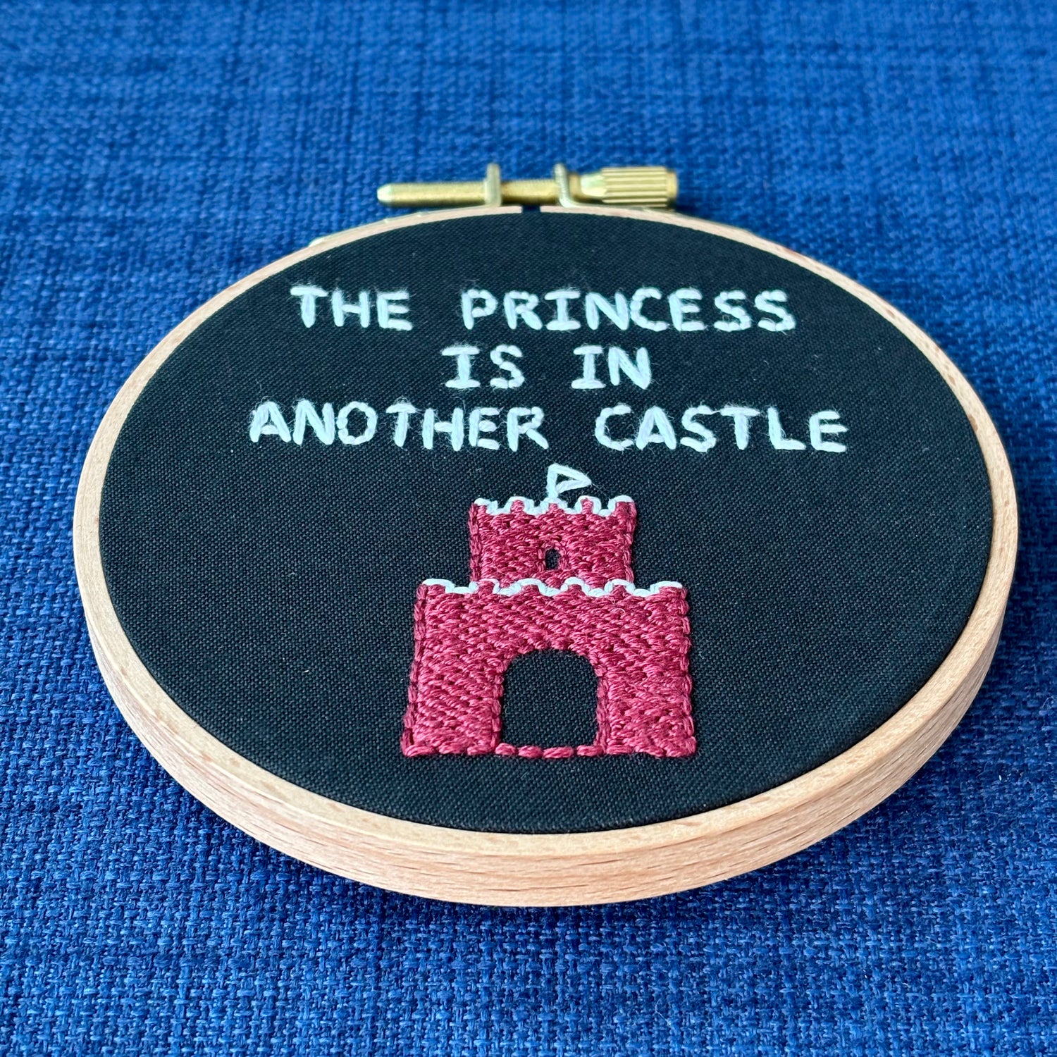 Embroidery hoop reads, "The princess is in another castle. Text is all capital letters in sans serif font and hand embroidered in white. Below the words is a brick castle with a triangular white flag on the top. Hand stitched on black cotton. Framed in light wood embroidery hoop and displayed as wall art or DIY handmade Christmas ornament.