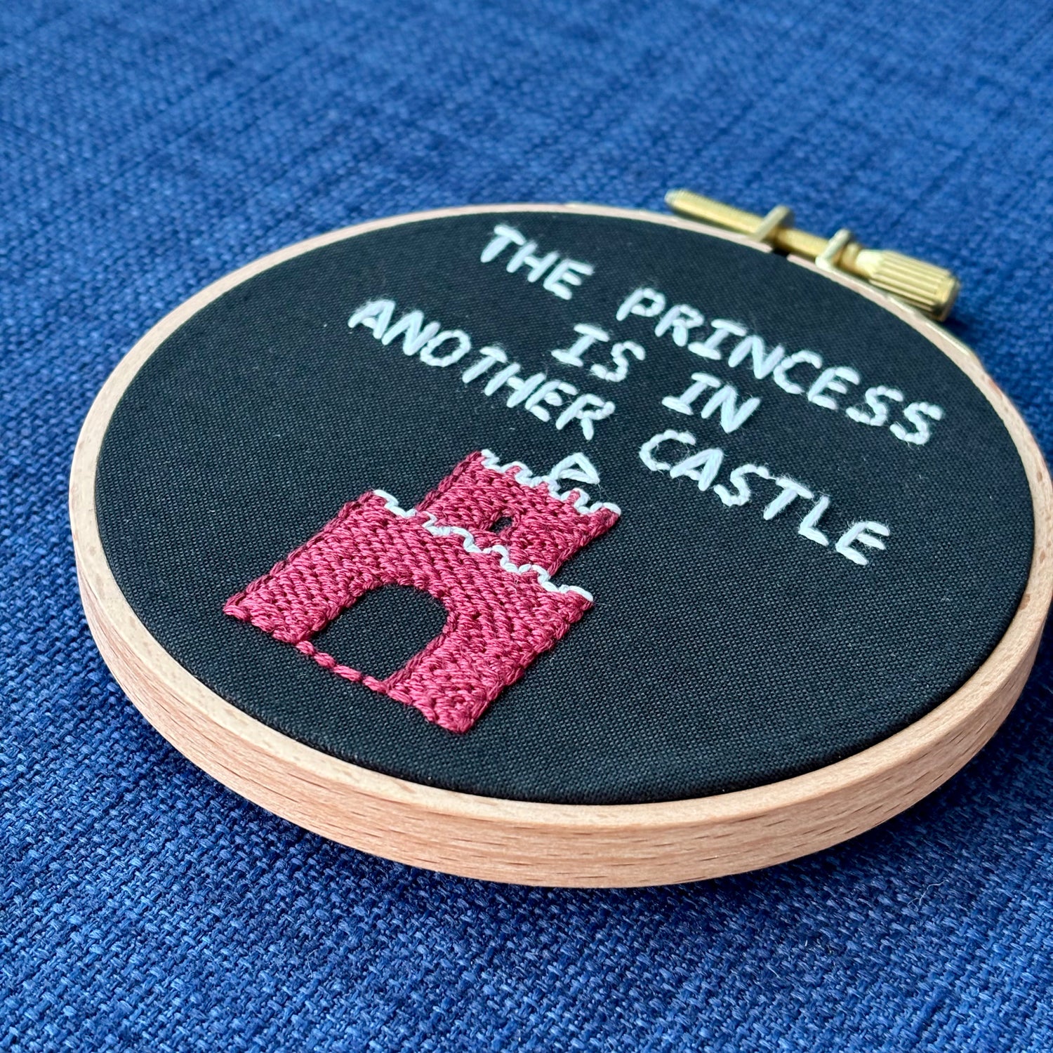 Embroidery hoop reads, "The princess is in another castle. Text is all capital letters in sans serif font and hand embroidered in white. Below the words is a brick castle with a triangular white flag on the top. Hand stitched on black cotton. Framed in light wood embroidery hoop and displayed as wall art or DIY handmade Christmas ornament.