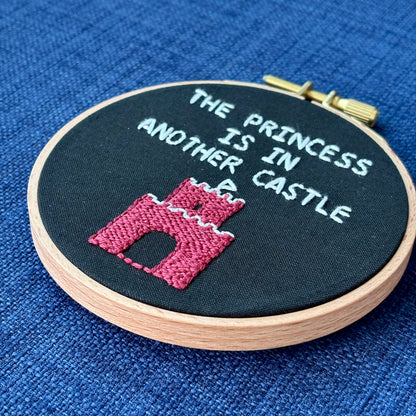 Embroidery hoop reads, "The princess is in another castle. Text is all capital letters in sans serif font and hand embroidered in white. Below the words is a brick castle with a triangular white flag on the top. Hand stitched on black cotton. Framed in light wood embroidery hoop and displayed as wall art or DIY handmade Christmas ornament.