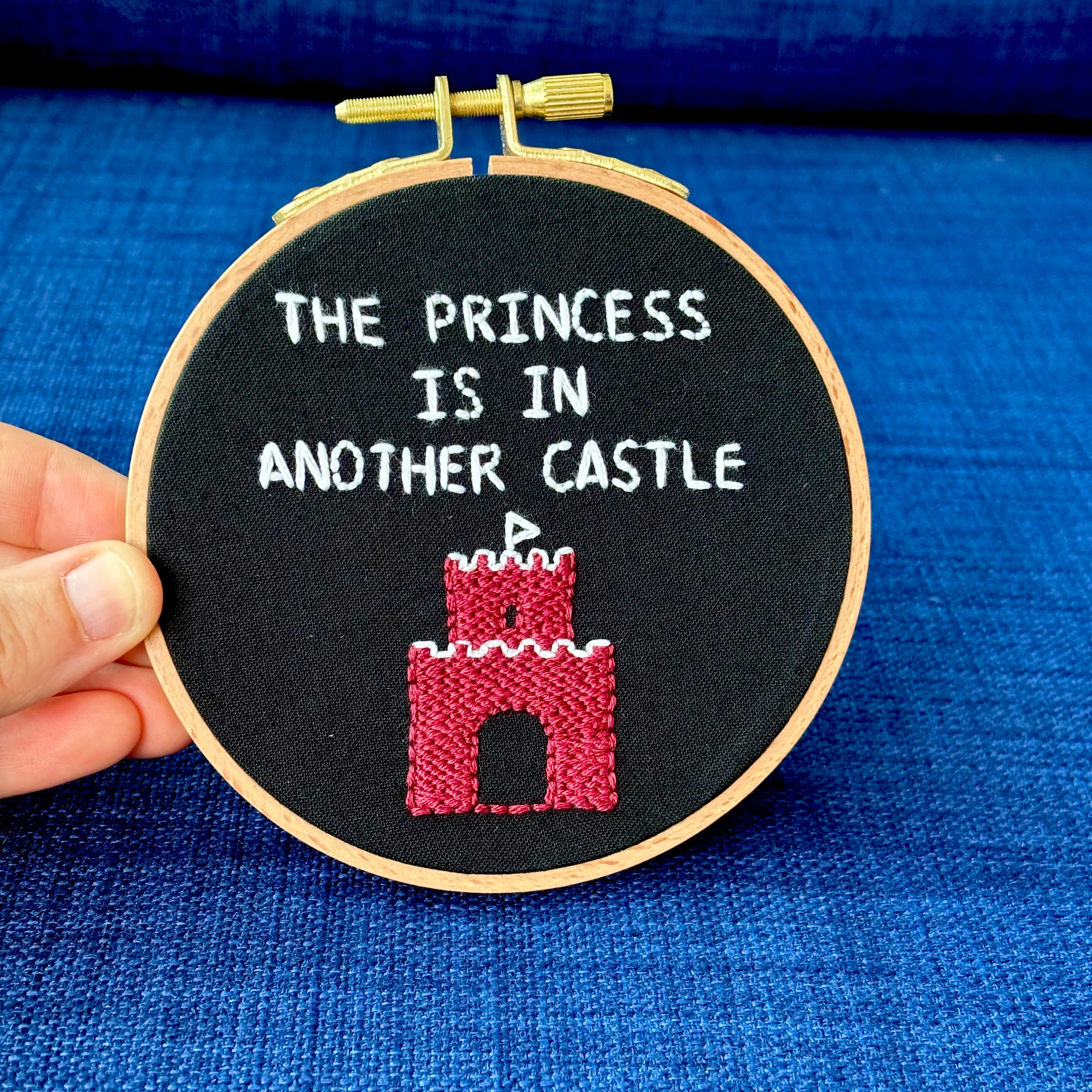 Embroidery hoop reads, "The princess is in another castle. Text is all capital letters in sans serif font and hand embroidered in white. Below the words is a brick castle with a triangular white flag on the top. Hand stitched on black cotton. Framed in light wood embroidery hoop and displayed as wall art or DIY handmade Christmas ornament.