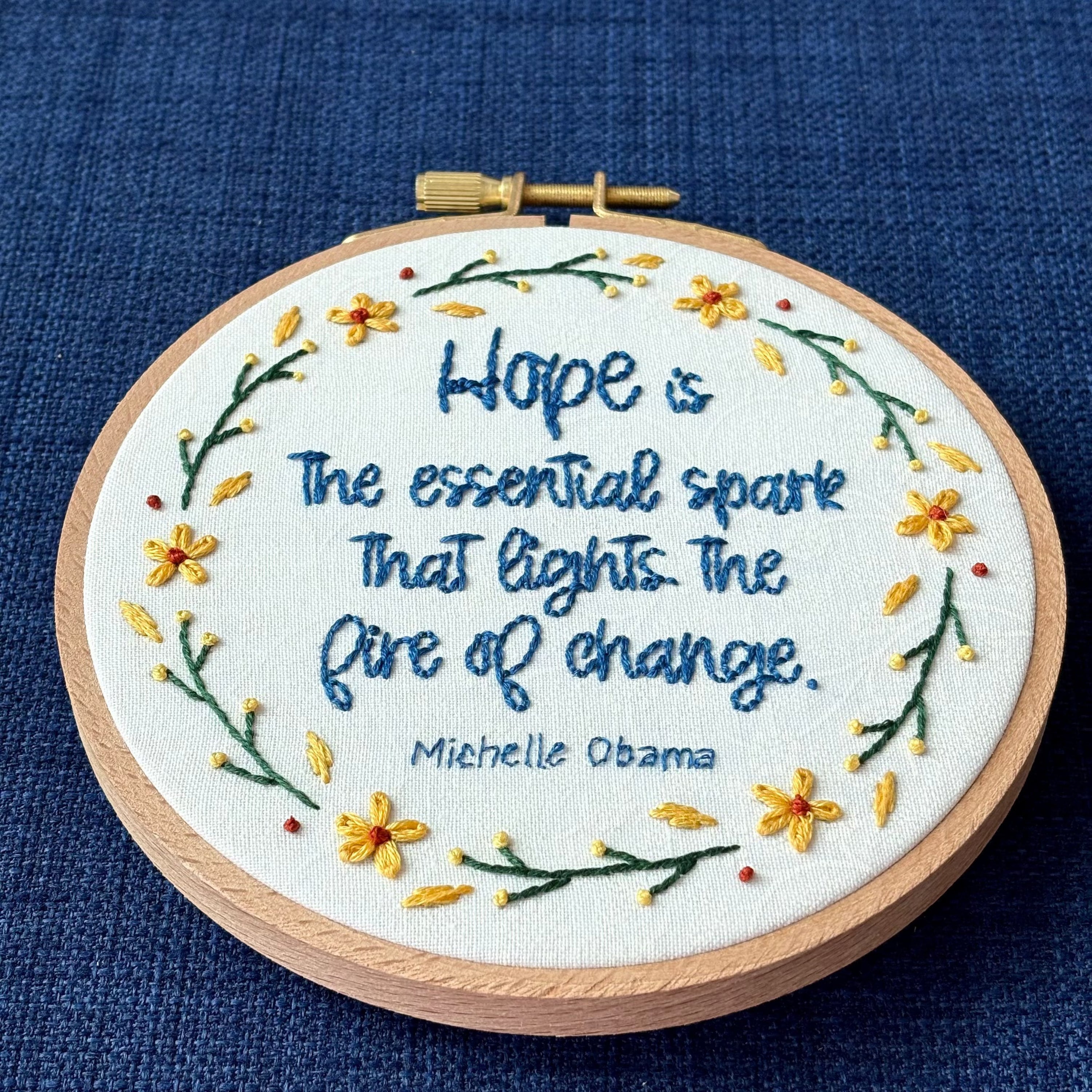 "Hope is the essential spark that lights the fire of change. Michelle Obama" Quote hand embroidered in dark blue. Text is surrounded by floral wreath of golden yellow sunflowers and dark green stems with light yellow buds. Fabric is white-on-white cotton. Displayed as wall art in a light wooden embroidery hoop.