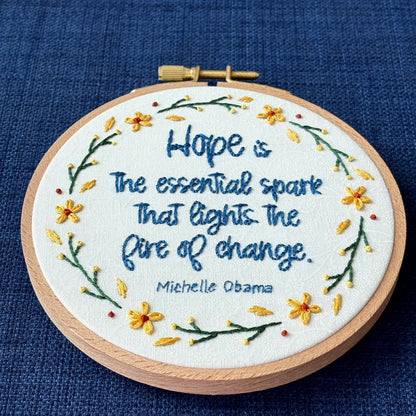 "Hope is the essential spark that lights the fire of change. Michelle Obama" Quote hand embroidered in dark blue. Text is surrounded by floral wreath of golden yellow sunflowers and dark green stems with light yellow buds. Fabric is white-on-white cotton. Displayed as wall art in a light wooden embroidery hoop.