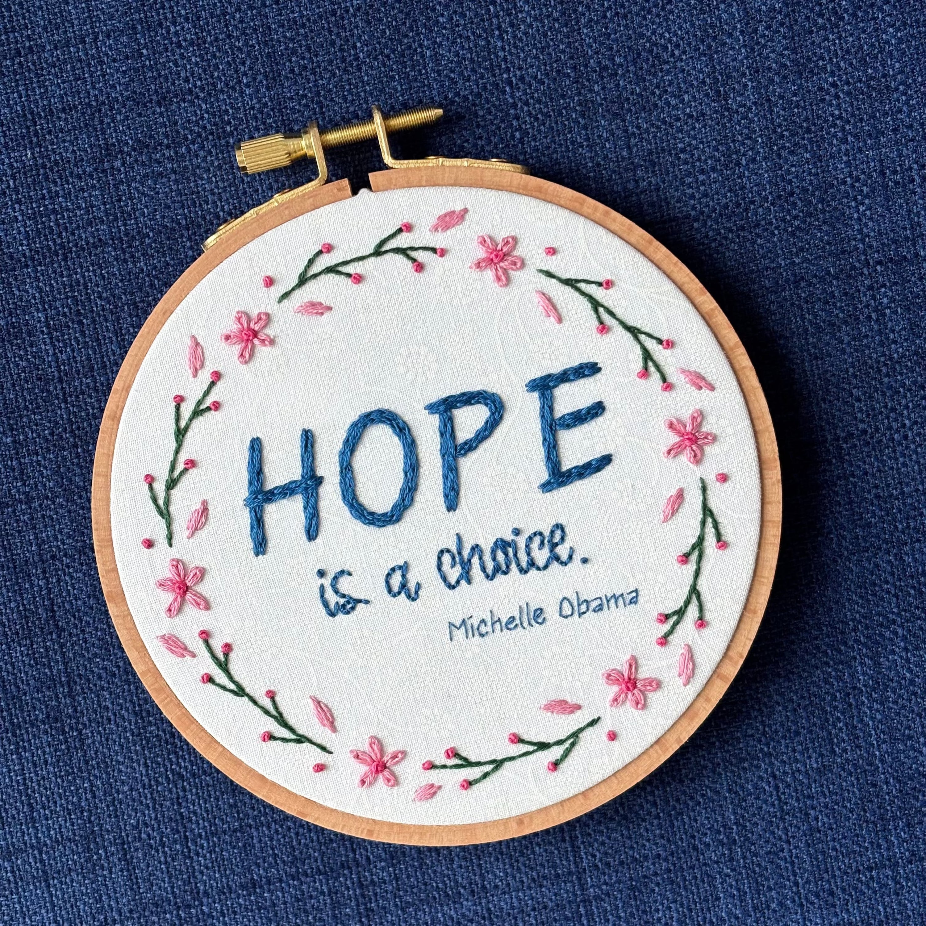 "Hope is a choice. Michelle Obama" Quote hand embroidered in dark blue. Text is surrounded by floral wreath of pink cherry blossom flowers and stems with buds. Fabric is white-on-white cotton. Displayed as wall art in a light wooden embroidery hoop.
