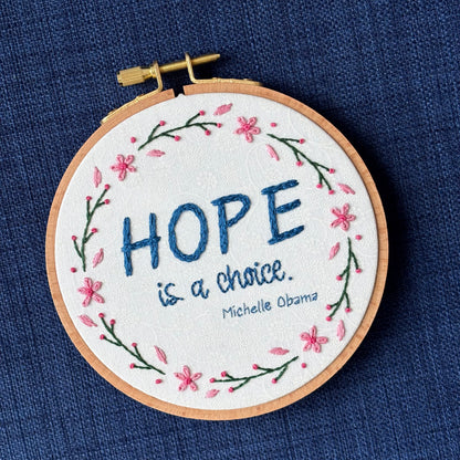"Hope is a choice. Michelle Obama" Quote hand embroidered in dark blue. Text is surrounded by floral wreath of pink cherry blossom flowers and stems with buds. Fabric is white-on-white cotton. Displayed as wall art in a light wooden embroidery hoop.