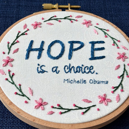 "Hope is a choice. Michelle Obama" Quote hand embroidered in dark blue. Text is surrounded by floral wreath of pink cherry blossom flowers and stems with buds. Fabric is white-on-white cotton. Displayed as wall art in a light wooden embroidery hoop.