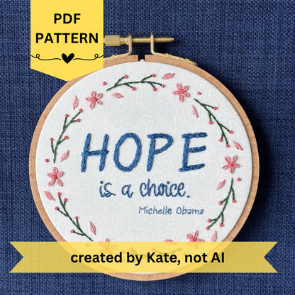 PDF pattern created by Kate, not AI. "Hope is a choice. Michelle Obama" Quote hand embroidered in dark blue. Text is surrounded by floral wreath of pink cherry blossom flowers and stems with buds. Fabric is white-on-white cotton. Displayed as wall art in a light wooden embroidery hoop.