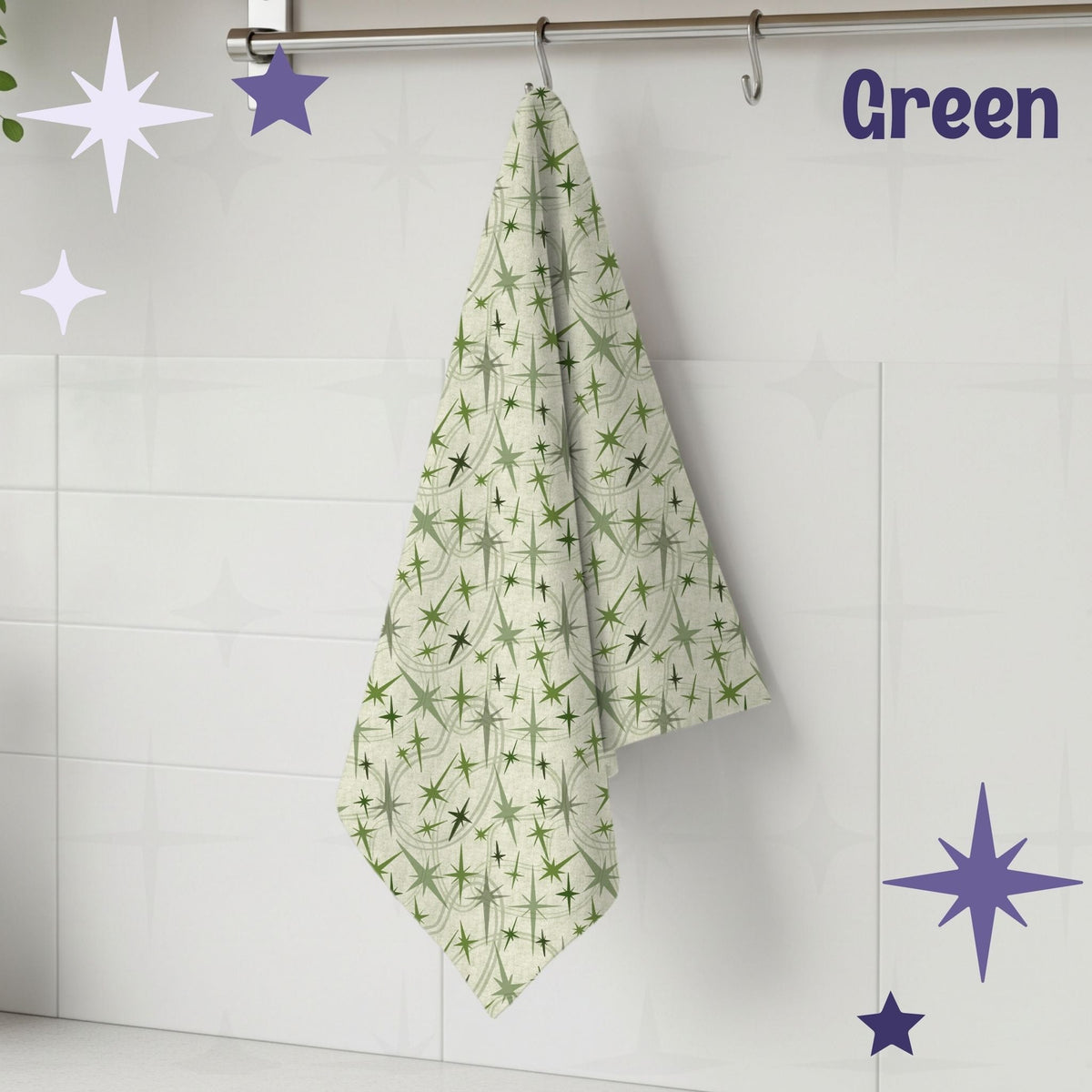 Sage green atomic starburst mid century modern tea towel hanging from a chrome hook against white subway tile, showing full pattern of olive and soft green starburst motifs on a warm cream cotton twill background