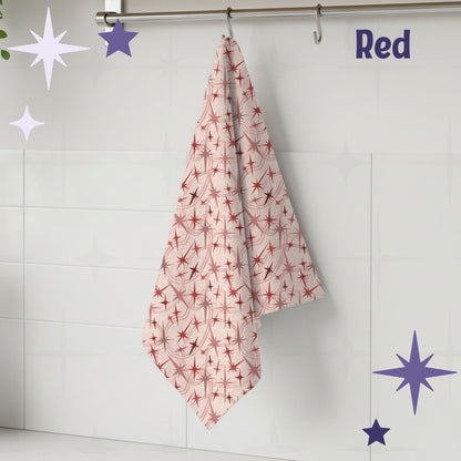 Red and dusty rose atomic starburst mid century modern tea towel hanging from a chrome hook against white subway tile, showing full pattern of crimson and blush starburst motifs on a nude cream cotton twill background