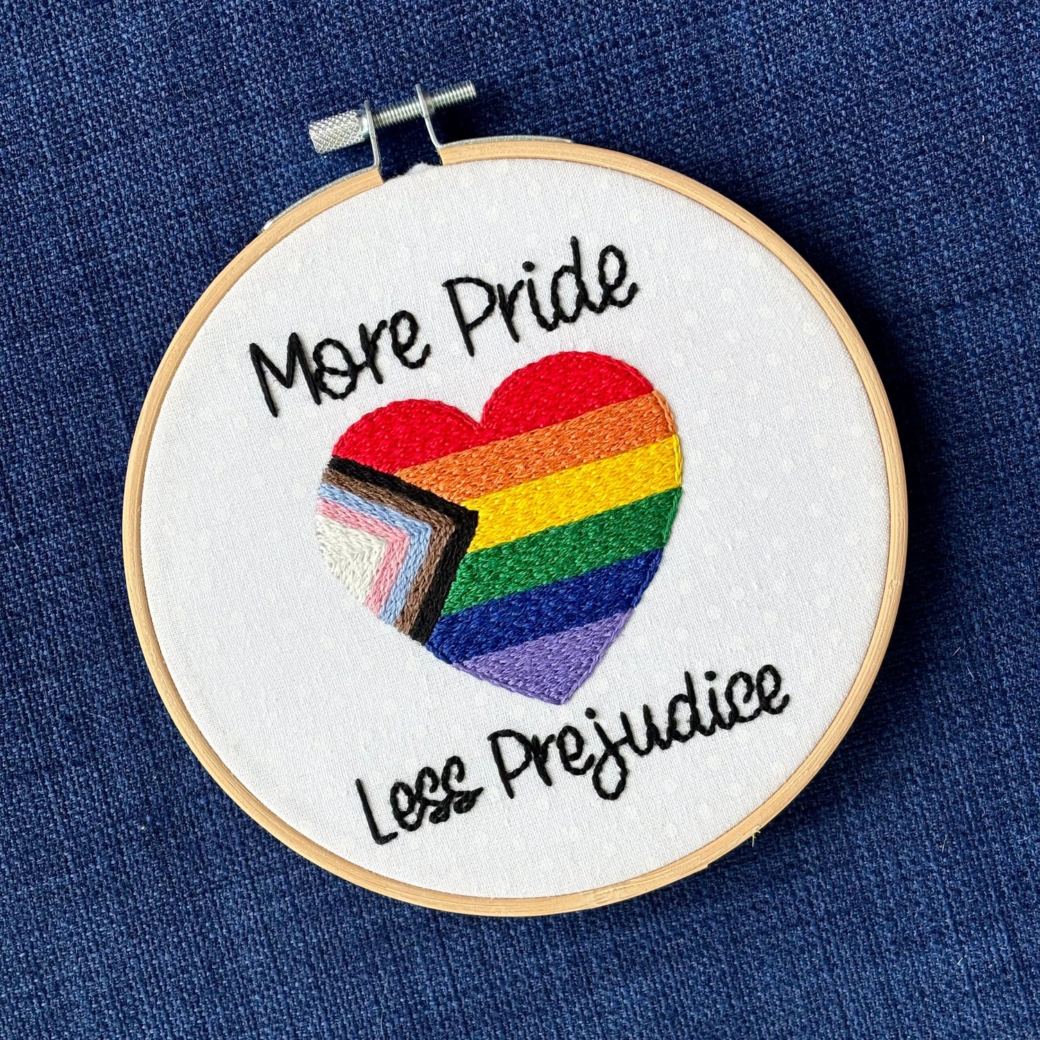 "More Pride Less Prejudice." Text in script font and hand embroidered in black. Words are placed above and below a heart shaped progress pride flag. Rainbow heart flag includes white, pink, light blue, brown, black, red, orange, yellow, green, blue, and purple stripes. Fabric is white cotton with white polka dots. Framed as wall art in light wood embroidery hoop.