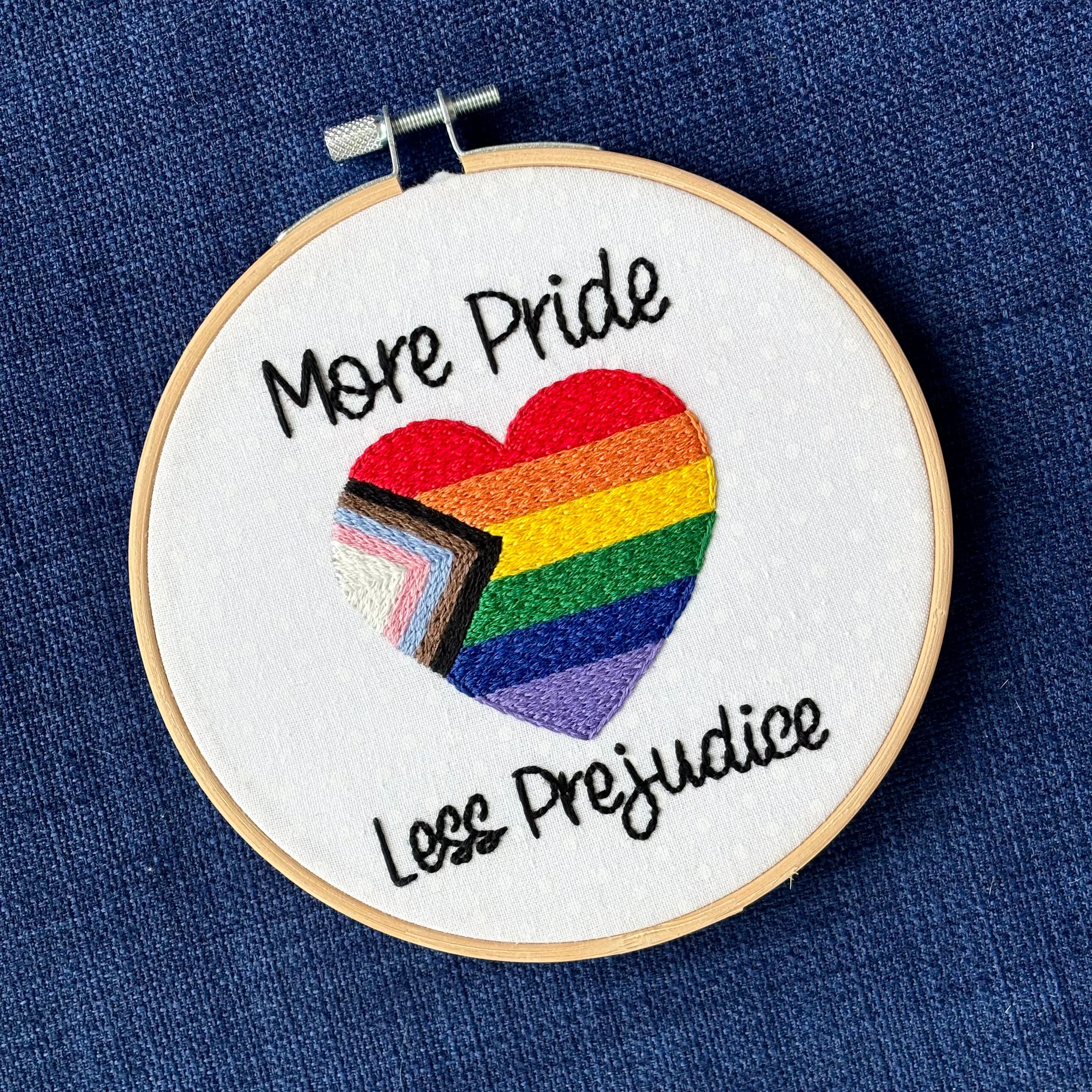 "More Pride Less Prejudice." Text in script font and hand embroidered in black. Words are placed above and below a heart shaped progress pride flag. Rainbow heart flag includes white, pink, light blue, brown, black, red, orange, yellow, green, blue, and purple stripes. Fabric is white cotton with white polka dots. Framed as wall art in light wood embroidery hoop.