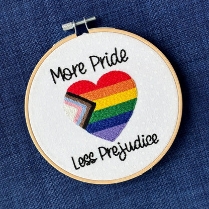 "More Pride Less Prejudice." Text in script font and hand embroidered in black. Words are placed above and below a heart shaped progress pride flag. Rainbow heart flag includes white, pink, light blue, brown, black, red, orange, yellow, green, blue, and purple stripes. Fabric is white cotton with white polka dots. Framed as wall art in light wood embroidery hoop.
