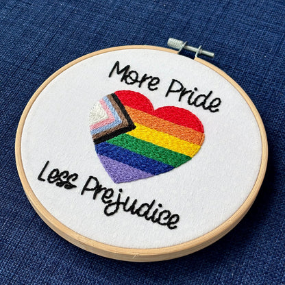"More Pride Less Prejudice." Text in script font and hand embroidered in black. Words are placed above and below a heart shaped progress pride flag. Rainbow heart flag includes white, pink, light blue, brown, black, red, orange, yellow, green, blue, and purple stripes. Fabric is white cotton with white polka dots. Framed as wall art in light wood embroidery hoop.