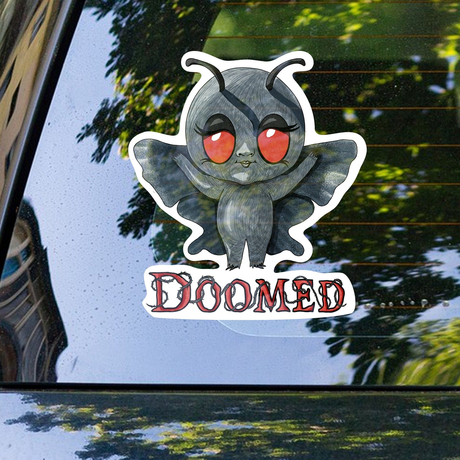 Mothman cryptid sticker on the rear window of a car with trees reflected behind
