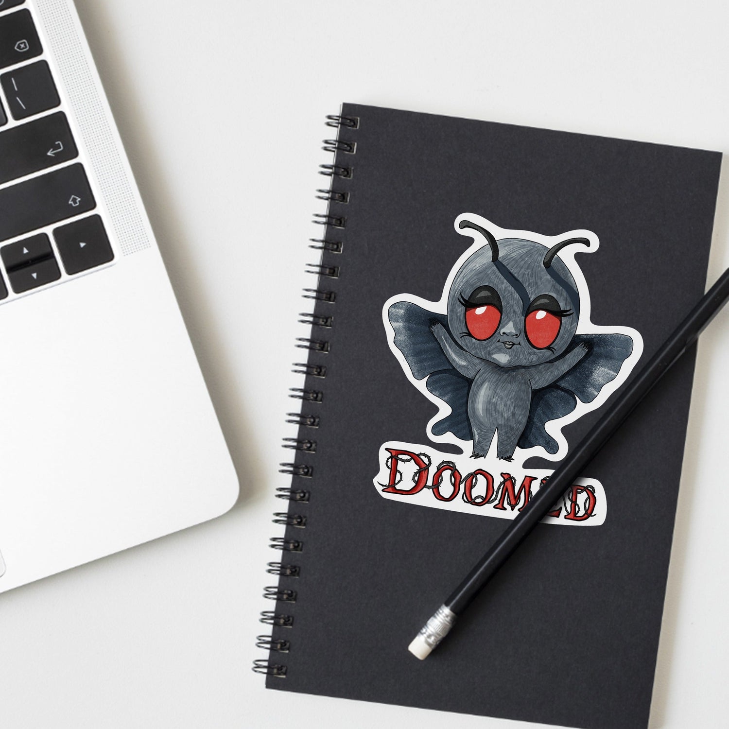 Chibi Mothman cryptid sticker on a black spiral notebook beside a laptop and pencil