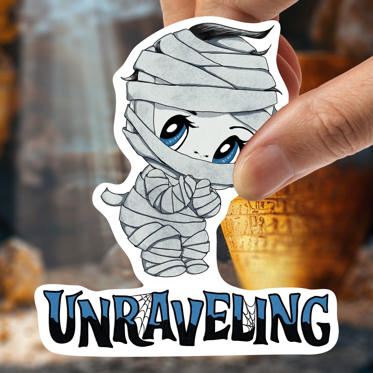 Hand holding the pastel goth mummy sticker against a golden ancient tomb background