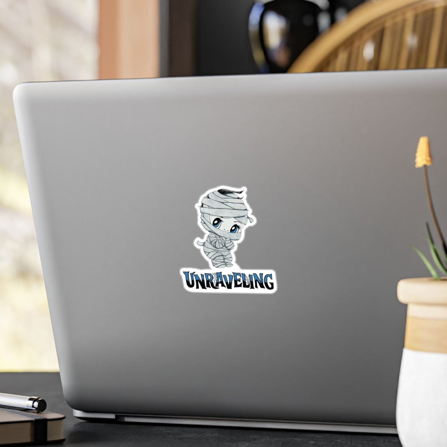 Small chibi mummy sticker with blue eyes and UNRAVELING lettering on the back of a silver laptop