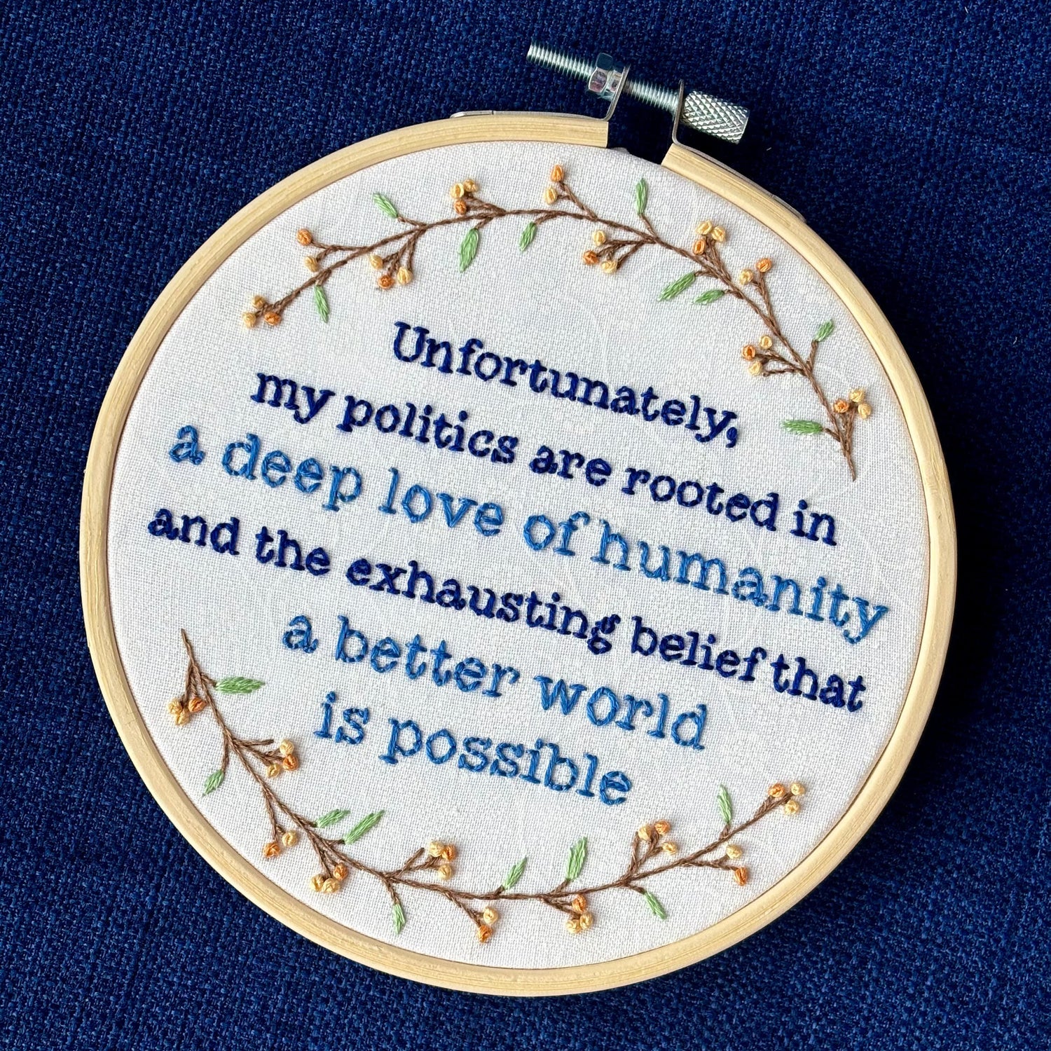 "Unfortunately, my politics are rooted in a deep love of humanity and the exhausting belief that a better world is possible." Quote hand embroidered in dark blue and medium blue. Text is surrounded by branches with light green leaves and light and medium pastel orange flower buds. Wall art is displayed in a light colored wood embroidery hoop.