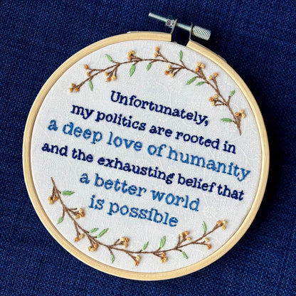 "Unfortunately, my politics are rooted in a deep love of humanity and the exhausting belief that a better world is possible." Quote hand embroidered in dark blue and medium blue. Text is surrounded by branches with light green leaves and light and medium pastel orange flower buds. Wall art is displayed in a light colored wood embroidery hoop.