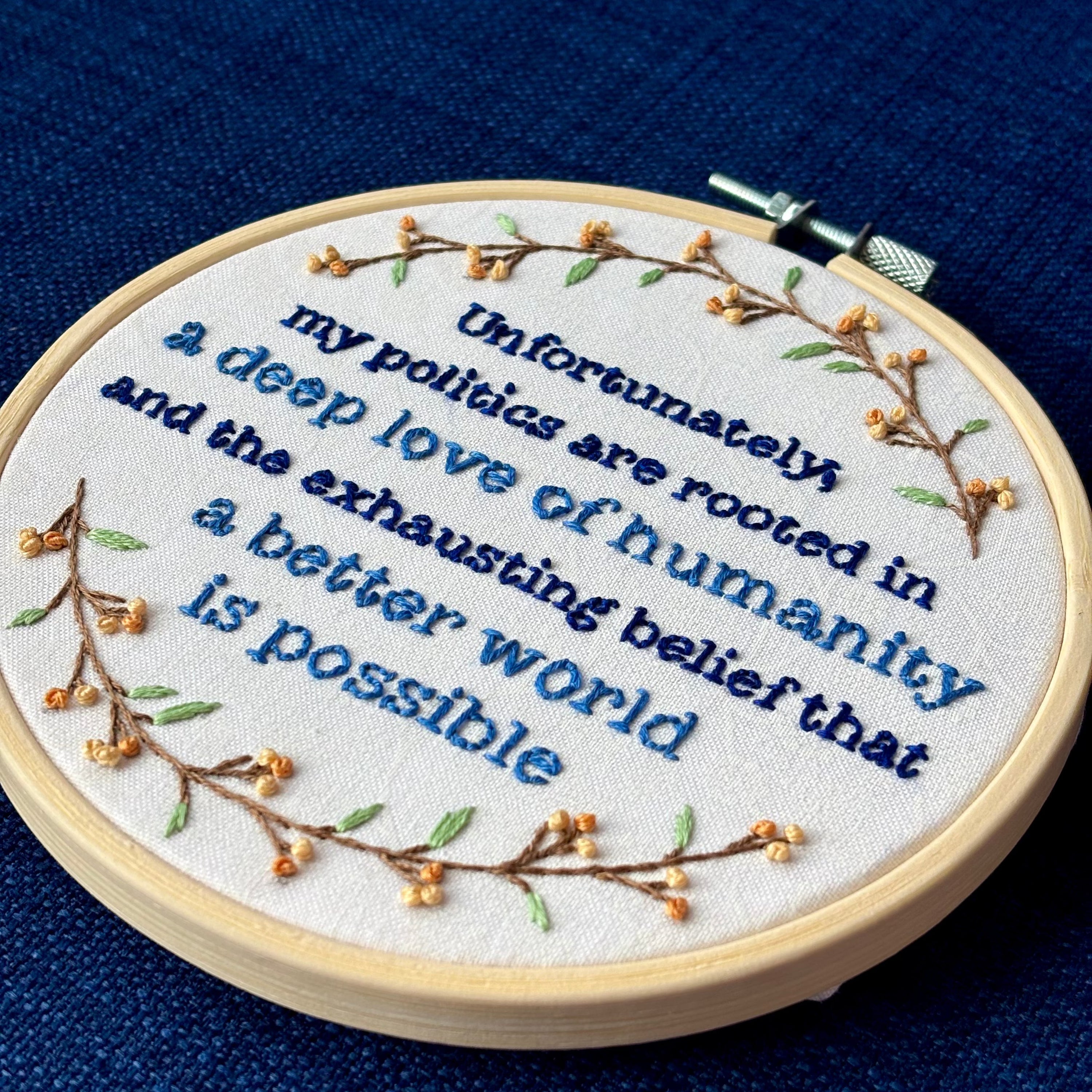 "Unfortunately, my politics are rooted in a deep love of humanity and the exhausting belief that a better world is possible." Quote hand embroidered in dark blue and medium blue. Text is surrounded by branches with light green leaves and light and medium pastel orange flower buds. Wall art is displayed in a light colored wood embroidery hoop.