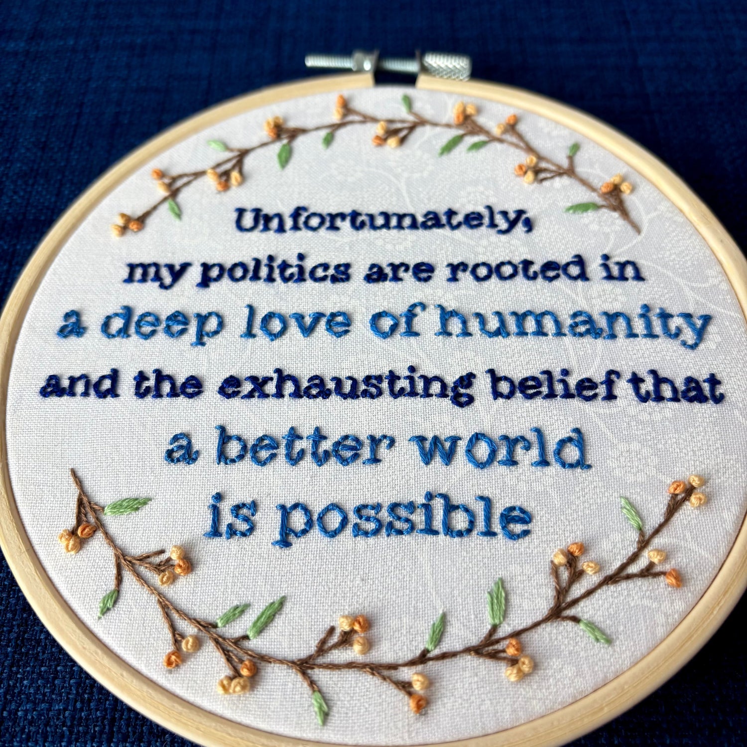 "Unfortunately, my politics are rooted in a deep love of humanity and the exhausting belief that a better world is possible." Quote hand embroidered in dark blue and medium blue. Text is surrounded by branches with light green leaves and light and medium pastel orange flower buds. Wall art is displayed in a light colored wood embroidery hoop.