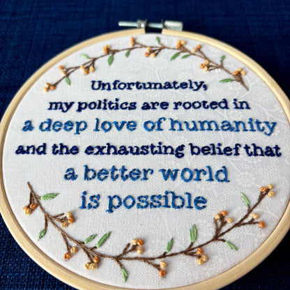 "Unfortunately, my politics are rooted in a deep love of humanity and the exhausting belief that a better world is possible." Quote hand embroidered in dark blue and medium blue. Text is surrounded by branches with light green leaves and light and medium pastel orange flower buds. Wall art is displayed in a light colored wood embroidery hoop.