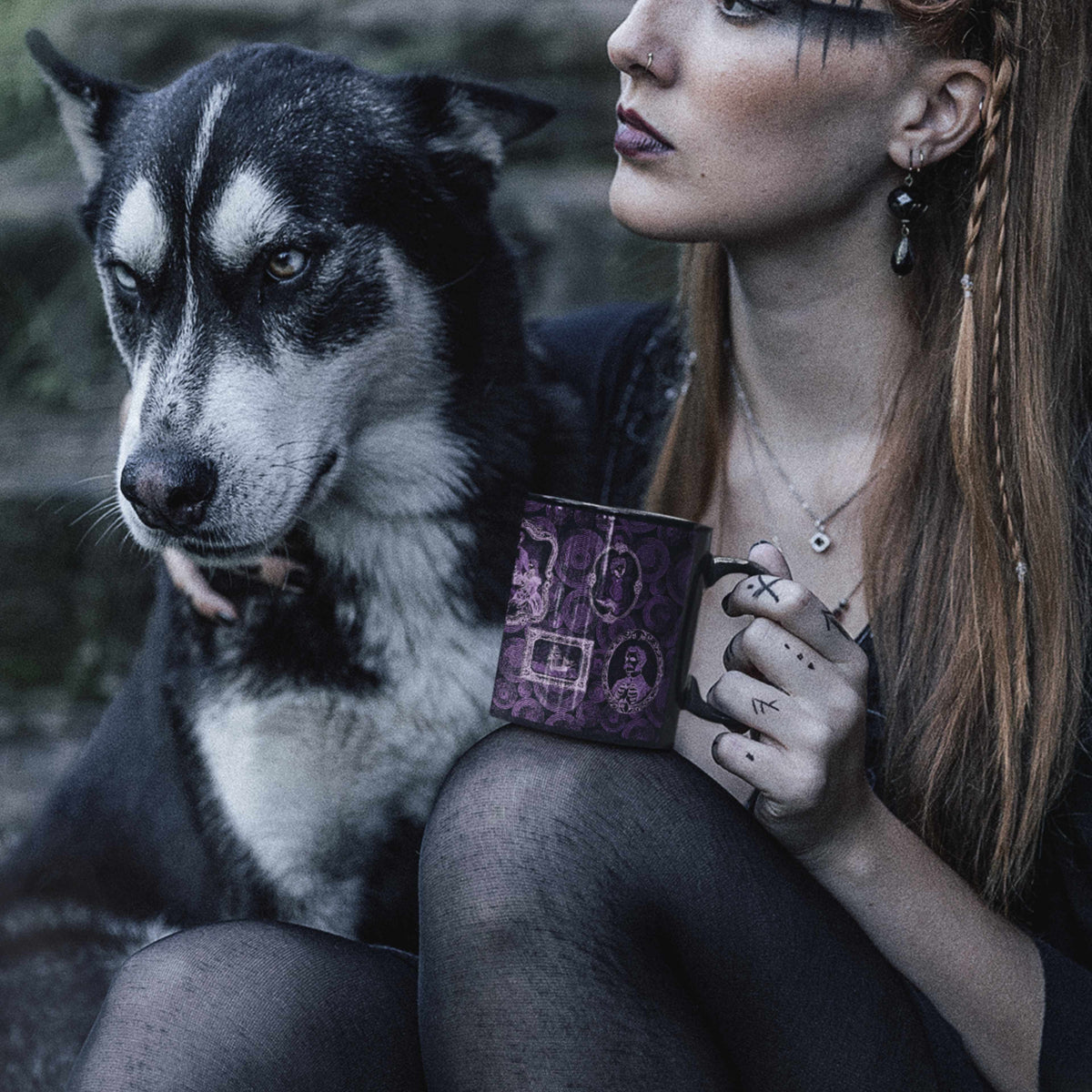 Woman with braided hair and rune tattoos holding a purple zombie portrait mug outdoors at dusk beside a husky