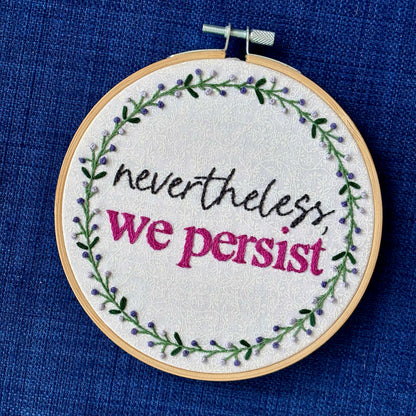 "Nevertheless, we persist." Nevertheless hand embroidered in a script font in dark gray. "we persist" is in Times New Roman and sewn bright fuchsia embroidery thread. Quote surrounded with a floral wreath with purple and lavender flower buds and green leaves. Stitched on white-on-white cotton. Wall art framed in light wood embroidery hoop.
