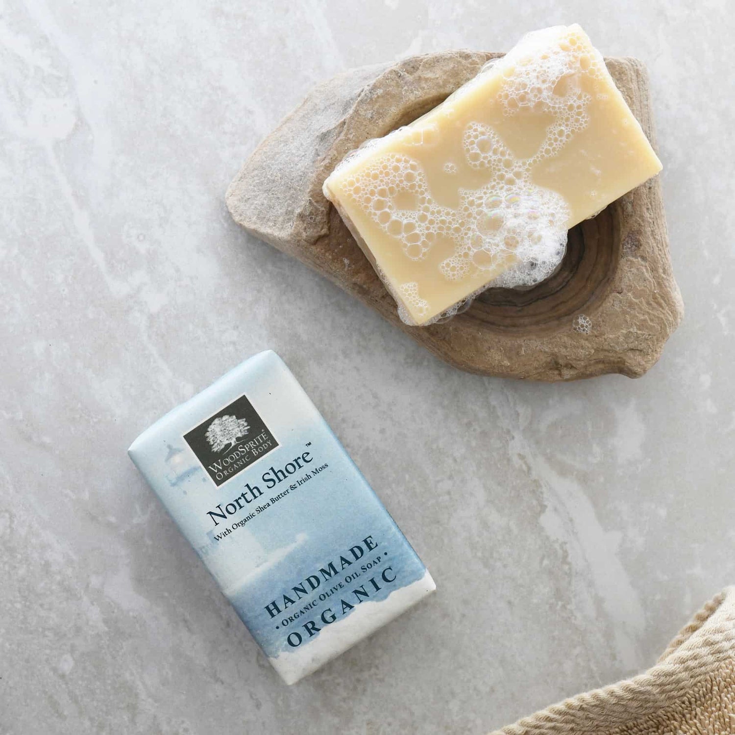 North Shore Organic Soap resting on a natural stone dish, lathered and sudsy, alongside its pale blue watercolor-printed packaging on a grey marble surface with a linen towel.