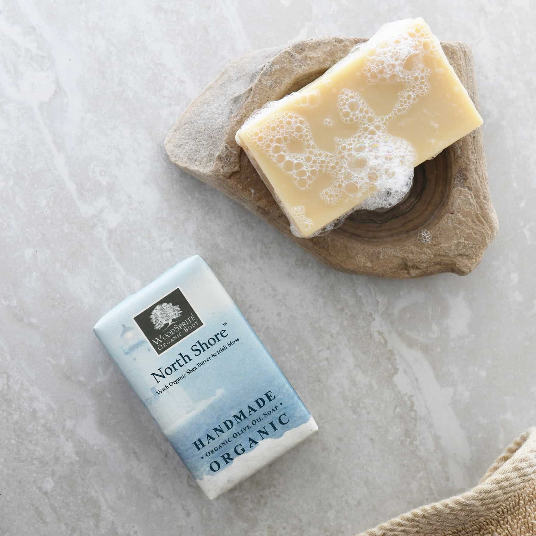 North Shore Organic Soap resting on a natural stone dish, lathered and sudsy, alongside its pale blue watercolor-printed packaging on a grey marble surface with a linen towel.