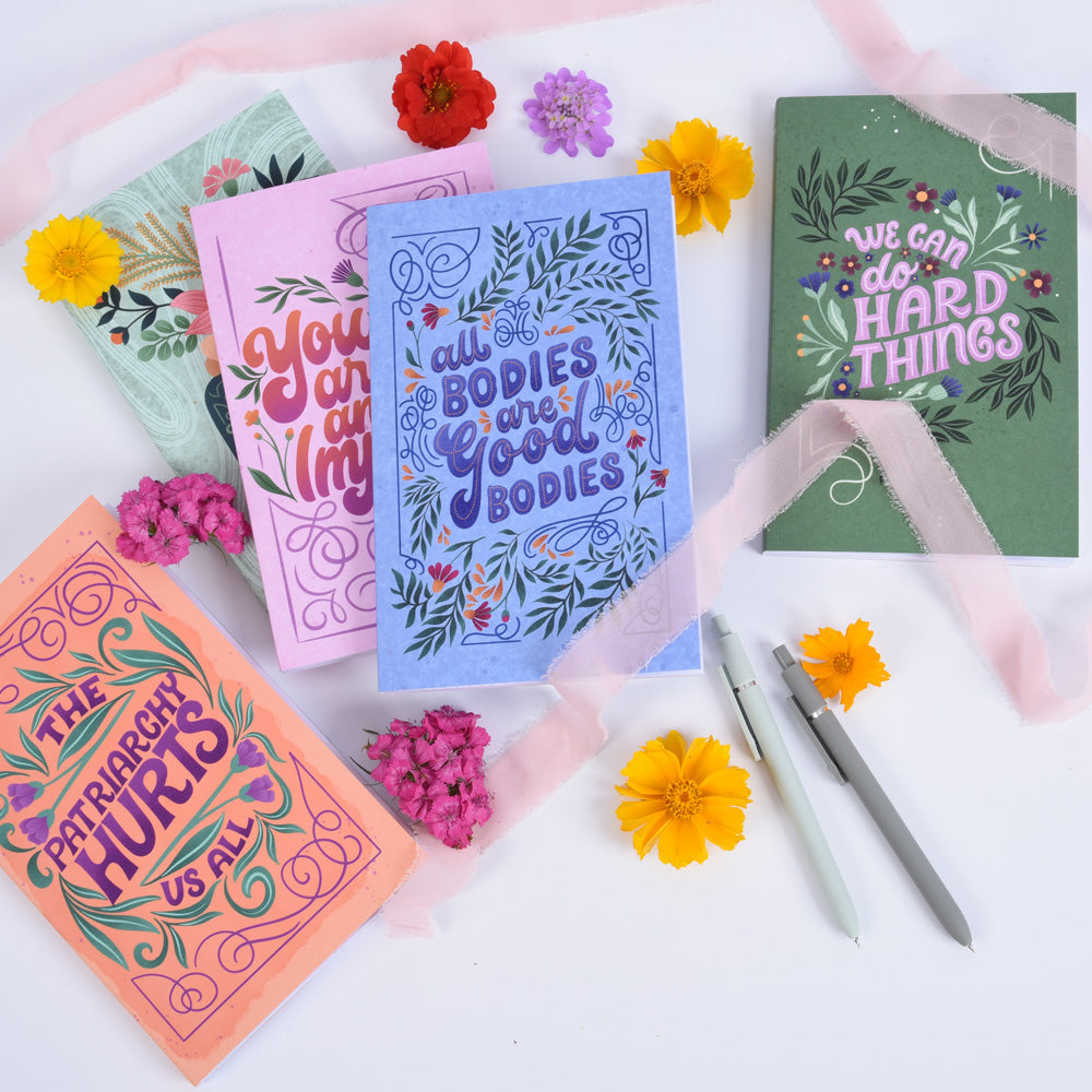 5 notebook journals with hand lettering and illustrated covers arranged on a white table with ribbons, pens, and colorful flowers