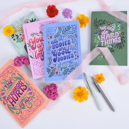 5 notebook journals with hand lettering and illustrated covers arranged on a white table with ribbons, pens, and colorful flowers