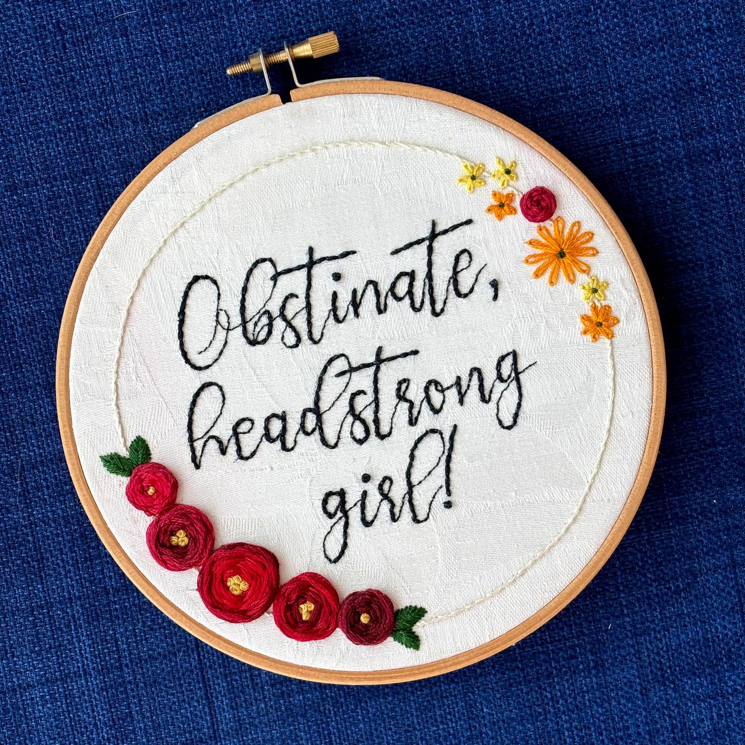 "Obstinate, headstrong girl!" Quote hand embroidered in sparkle black thread surrounded by a floral wreath with red roses and yellow and orange daisies. Fabric is white damask. Embroidery is displayed as wall art in light wood embroidery hoop.